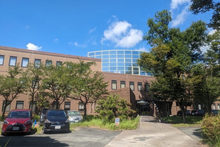 晴れた日の会社外観。レンガ色の3階建て建物で、上部に円形のガラス張りの構造物がある。前面に駐車場があり、数台の車が停まっている。建物の周りには緑豊かな木々が植えられ、青空と白い雲が見える。