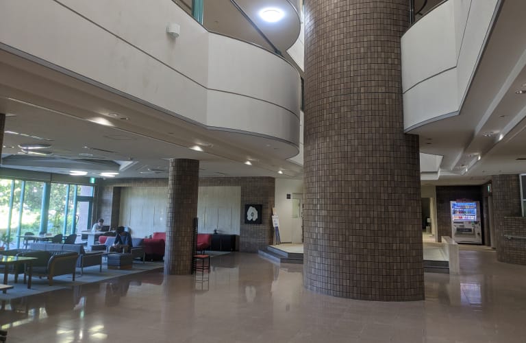 広々とした会社のエントランスホール。大きな円柱と曲線的な2階部分が特徴的。左側にはロビーエリアがあり、ソファや椅子が配置されている。床は光沢のある白い床材。