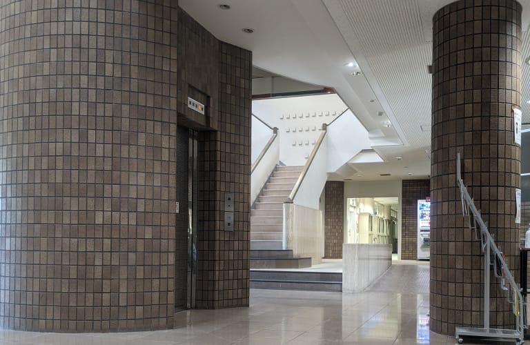 会社の内部。タイル張りの円柱とエレベーター、奥に階段が見える。床は光沢のある白い床材で、天井は格子状のデザイン。