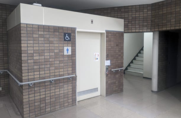 車椅子用トイレ入口。茶色のタイル壁に白いドア、車椅子のシンボルマークが掲示されている。右側に階段が見える。