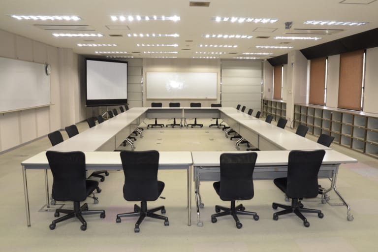 大型の貸会議室の内部。四角に配置された白いテーブルと黒い椅子、正面にはスクリーンとホワイトボード、天井には明るい照明が並ぶ。壁際には棚が設置され、窓にはブラインドが掛かっている。